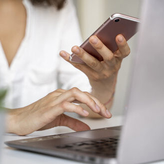 close-up-of-woman-using-cell-phone-and-laptop-at-desk-AFVF03714